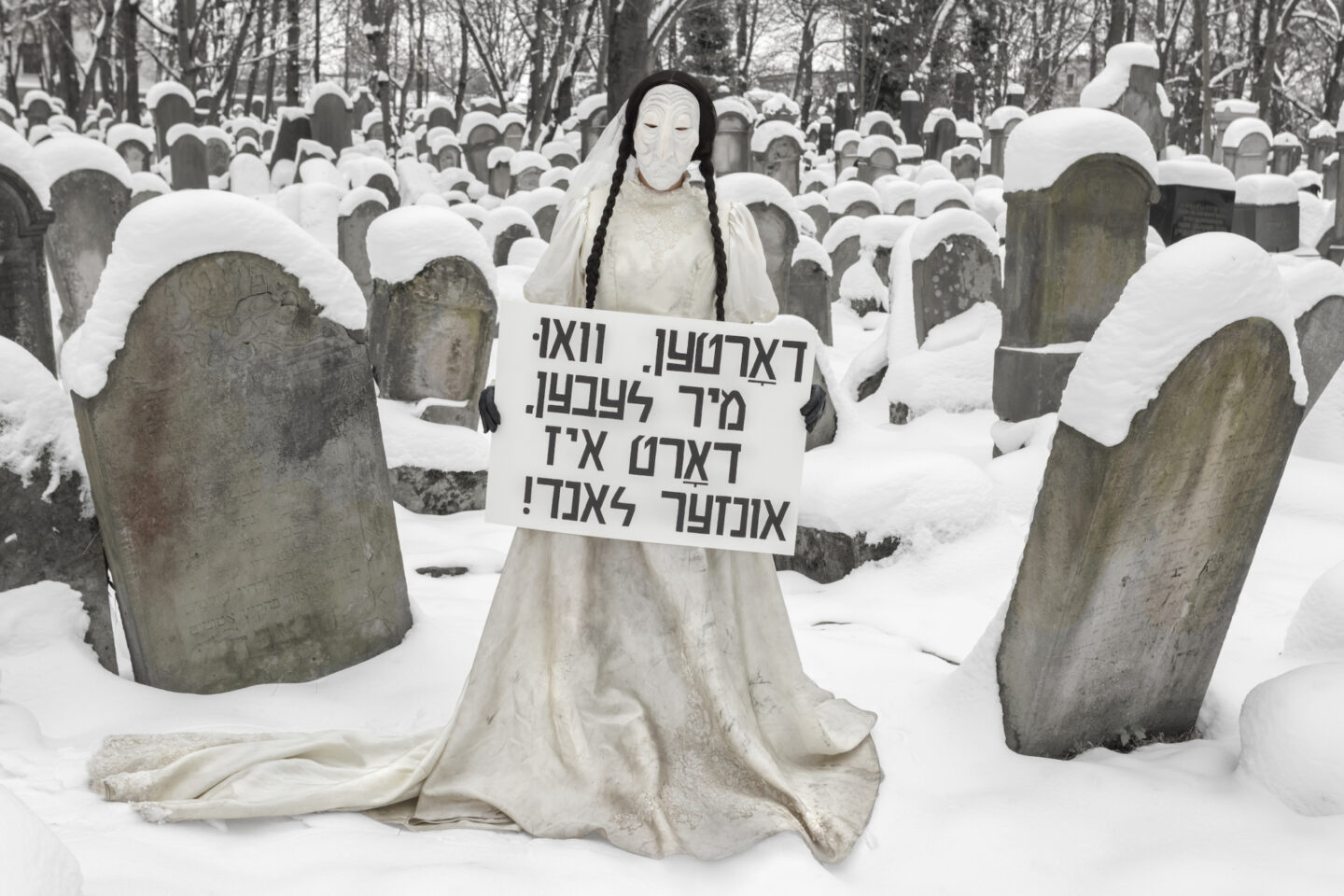 <p dir="ltr">Julie Weitz, <em>Wherever We Live That’s Our Homeland (Dybbuk in Jewish Cemetery, Tarnow, Poland)</em> 2024 (photo credit Magda Chudzik)</p>
<p dir="ltr"><em>Image Description. Julie Weitz as her dybbuk character: She’s wearing a white dress, two long black braids, and a white veil, evocative of Leah’s costume in</em> Der Dybbuk<em>. She’s also wearing a white mask of a wrinkly, elderly face. She stands in a snow-covered Jewish cemetery, and holds a sign, which reads in Yiddish the Bundist slogan: “Dortn, vu mir lebn, dort iz undzer land”, meaning “Wherever we live, that is our homeland”.</em></p>
<p><br></p>