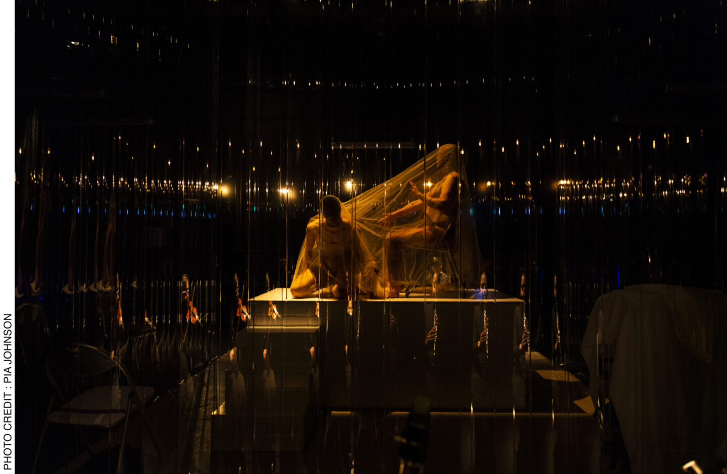 <p>Performers Lauren Langlois and Yoni Prior in the opening Act of <i>Dybbuks </i>are shrouded atop the central raised platform of the stage, moving slowly around each other’s bodies. The musicians’ reflections can be seen against the perspex curtain. Photo Credit: Pia Johnson.</p>