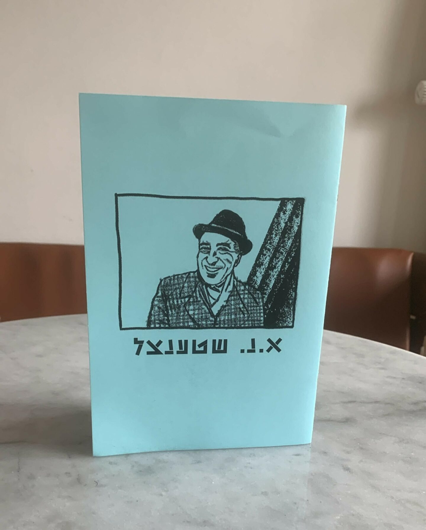 <p>A copy of the A.N. Stencl zine, <em>Geklibene lider/Selected Poems, </em>edited by Jake Schneider, with translations by Arndt Beck, Horst Bernhardt, Guli Dolev-Hashiloni, Jordan Lee Schnee, and Jake Schneider, published by Yiddish.Berlin in 2023. Illustration by Miriam Jacquiot. Photo courtesy of Jordan Lee Schnee. </p>