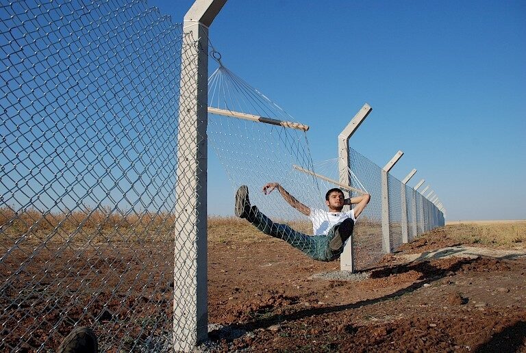 <p>Image: Documentation of a performance by Murat Gök titled “Border (Hammock),” 2010. The artist installed a hammock on the border fence separating Turkey, Syria, and the Kurdish territories, playfully satirizing the absolutism of borders. <a href="https://www.instituteforpublicart.org/case-studies/border/">https://www.instituteforpublicart.org/case-studies/border/</a></p>