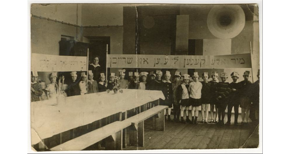 <p>Stefania Shifra Szabad, wife of Jewish communal leader Tsemaḥ Szabad, with a class in a Yiddish school, Vilna, 1929. Children, wearing hats representing letters of the <i>alefbeys</i> (alphabet), hold a banner (right) that reads in Yiddish: “We can all read and write.” (YIVO)</p>